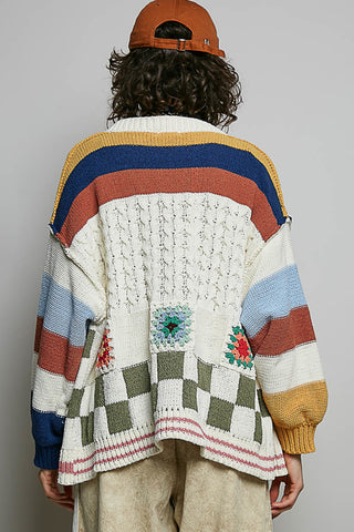 A person shown from behind wearing a chunky knit cardigan with wide multicolor stripes and patchwork details, paired with beige pants and an orange cap against a plain light gray background.