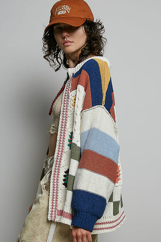 A model wearing a colorful striped knit cardigan with geometric patterns, paired with a brown cap and beige pants, photographed against a neutral gray background.