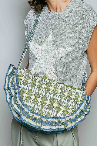 A person wearing a gray knit top with a large white star and carrying a ruffled shoulder bag with a blue and green botanical pattern on a white background.