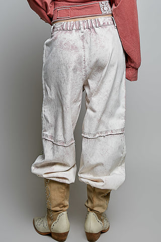 A person shown from the back wearing light-wash, heavily faded denim harem-style pants with a ruffled waistband and cuffed hems, paired with tan cowboy boots and a dusty rose top against a plain gray studio background.