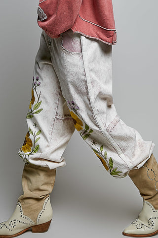 A close-up of a person's lower body wearing distressed off-white denim pants with yellow and green floral embroidery, paired with beige western-style boots and a dusty pink top, set against a plain gray studio background.