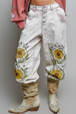 A person wearing distressed white denim pants with large yellow floral embroidery on the lower legs, styled with tan cowboy boots and a pink shirt against a plain gray studio background.
