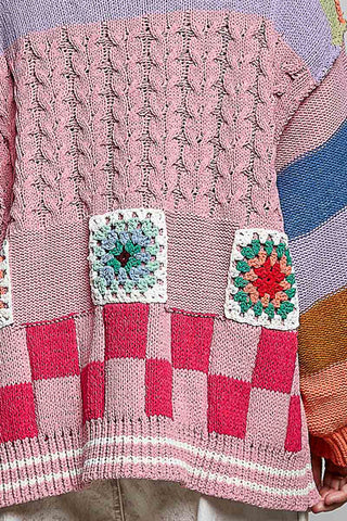 A close-up of a pink knitted sweater featuring textured cable-knit panels, a pink and red checkered lower section, and two colorful crocheted granny square patches on the front.