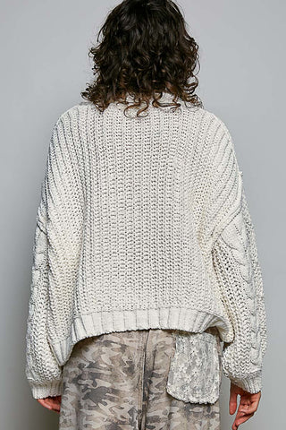 A person shown from the back wearing an oversized chunky knit cream sweater and camouflage pants against a plain light gray studio background.