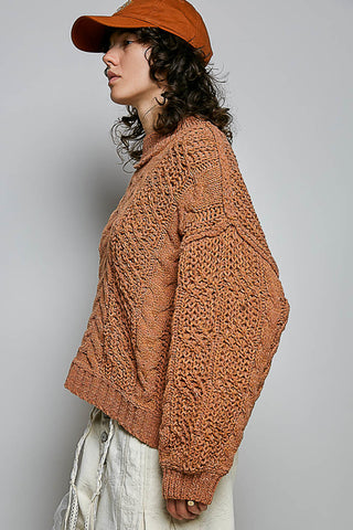 A woman shown in profile wearing a chunky, textured knit cardigan in a warm rust-orange color, paired with a matching orange cap and off-white pants, against a plain gray studio background.