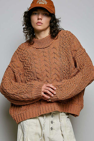 Person wearing a brown cable knit sweater and cap against a plain background