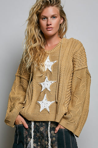 A woman wearing a chunky knit tan sweater with three large white star appliqués down the front, paired with dark patterned pants, against a plain gray studio background.