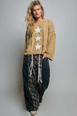 A woman modeling a tan cable-knit sweater with three large white star appliqués down the front, paired with dark patterned wide-leg pants, against a plain gray studio background.