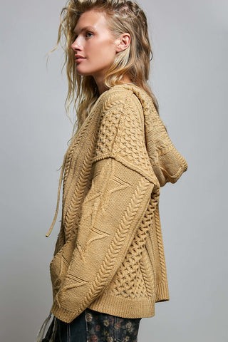 A woman wearing a chunky cable-knit hooded cardigan in a warm camel tan color, shown from a side profile against a solid gray studio background.