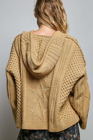 A woman viewed from behind wearing a chunky tan cable-knit hooded sweater with star patterns on the back, paired with dark floral pants, against a plain gray studio background.