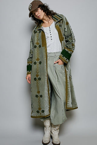 A person modeling a long, wide coat with embroidered floral and geometric patterns in olive green, brown, and cream, worn over a white top and matching wide-leg pants, against a plain light gray studio background.