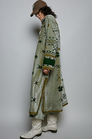 A model wearing a long sage green velvet coat with intricate green, gold, and brown embroidered patterns, paired with white western boots and a brown checkered cap, photographed against a neutral gray studio background.