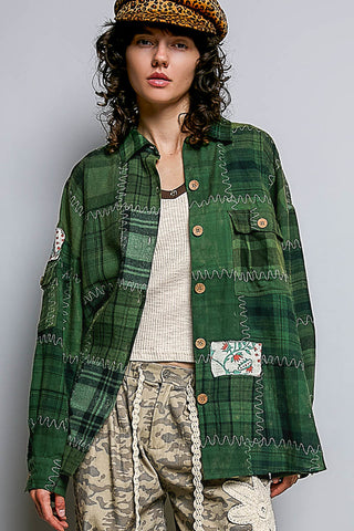 A model wearing a green plaid patchwork button-up shirt with zigzag stitching, a leopard print flat cap, a cream ribbed top, and camouflage pants against a solid gray background.