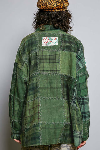 A person shown from behind wearing an oversized green patchwork plaid shirt with a floral applique patch on the back, a leopard print beret, and camouflage pants against a plain gray background.