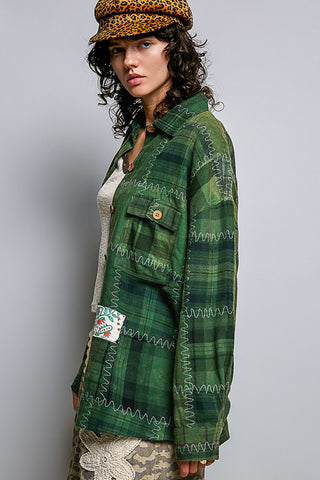 A woman wearing a green plaid button-up shirt jacket with white zigzag stitched details, a leopard print cap, and a white top, photographed against a plain gray studio background.