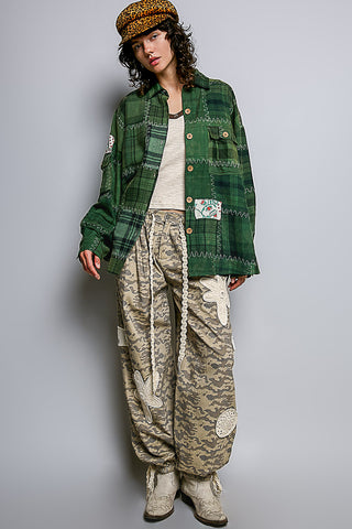 A model wearing a green plaid button-up shirt jacket, beige camouflage cargo pants, a patterned brown beret, and beige boots, standing against a light gray studio background.