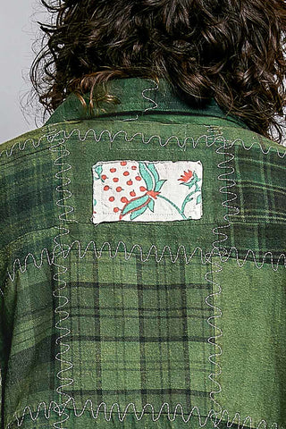 Close-up back view of a person wearing a green plaid flannel shirt with decorative zigzag stitching and a square floral patch on the upper back.