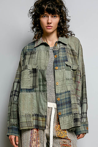 A model wearing a patchwork button-up shirt jacket made of olive green, blue, and brown plaid fabrics, styled with a gray t-shirt and patterned pants against a light gray studio background.