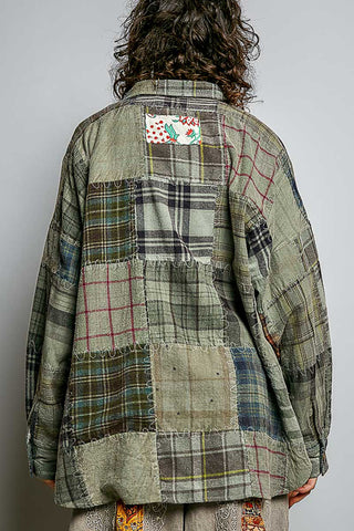 A person shown from the back wearing an oversized patchwork plaid shirt jacket in muted earthy tones of green, beige, gray, and brown, with a small colorful patch on the upper back.