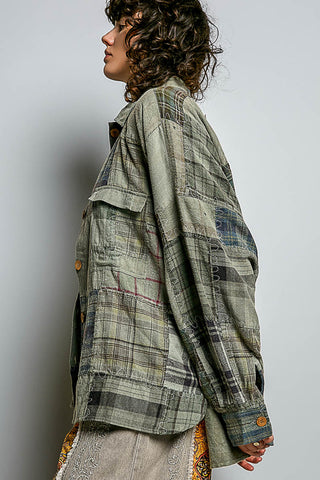 A model shown in profile wearing a long-sleeve patchwork plaid shirt jacket in muted green, gray, and beige tones, paired with a patterned skirt, against a plain light gray studio background.