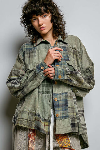 A model with curly dark hair wearing an oversized, patchwork-style plaid button-up shirt in muted green, blue, and beige tones, posed against a neutral gray studio background.