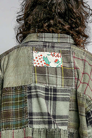Close-up back view of a person wearing a patchwork quilted jacket made of various plaid and patterned fabric panels, featuring a small colorful floral patch on the upper back.
