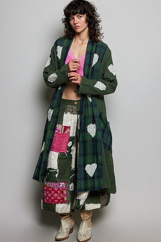 A model wearing a long green plaid patchwork cardigan with white heart patches, a pink top, and beige lace-up boots, standing against a plain gray studio background.