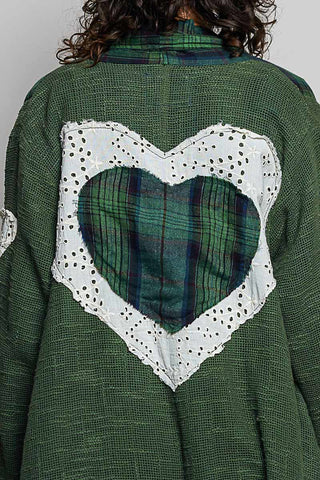 A close-up of the back of a green knit cardigan featuring a large heart-shaped patchwork design with white eyelet lace and a green plaid fabric insert.
