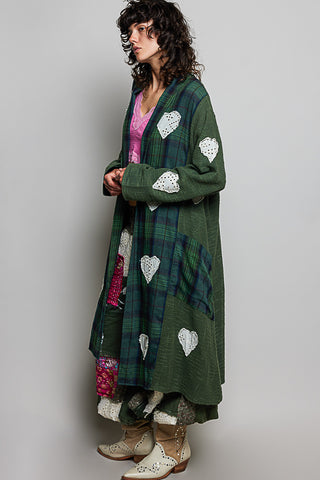 A woman models a long green plaid cardigan with white heart-shaped patches, worn over a pink top and patterned pants, against a plain gray studio background.