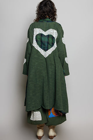 A person shown from the back wearing a long dark green textured coat with a large white crocheted heart outline and a green plaid heart insert on the back, paired with patterned green pants and beige shoes, against a plain light gray background.