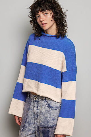 A model with curly dark hair wearing a blue and cream striped cropped knit sweater and acid-wash jeans, photographed against a neutral gray studio background.