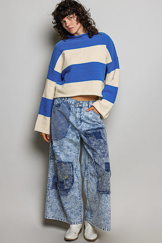 A model wearing a blue and cream striped knit sweater and wide-leg patchwork denim jeans, standing against a neutral gray studio background.