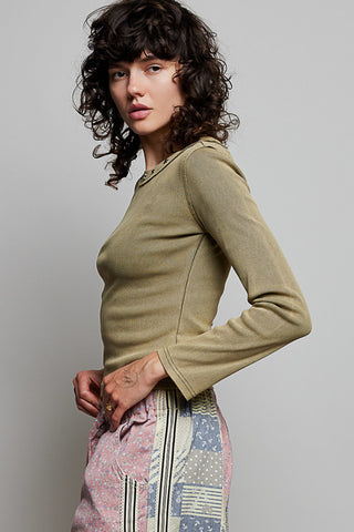 A model with curly dark hair is shown from the side, wearing a fitted, long-sleeve olive green top and patterned pink, blue, and white pants against a plain gray studio background.