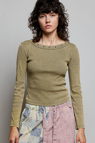 A model with curly dark hair wearing a ribbed olive green long-sleeve top with metal eyelets around the boat neckline, paired with patterned pink and blue pants, against a solid gray background.