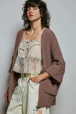 A woman wearing a chunky brown knit cardigan over a cream lace camisole and cream floral pants, photographed against a plain light gray studio background.