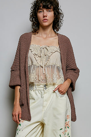 A woman wearing a chunky brown knit cardigan over a cream lace camisole and cream floral pants, photographed against a plain light gray studio background.