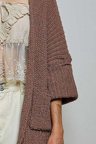 A woman wearing a chunky brown knit cardigan over a cream lace camisole and cream floral pants, photographed against a plain light gray studio background.