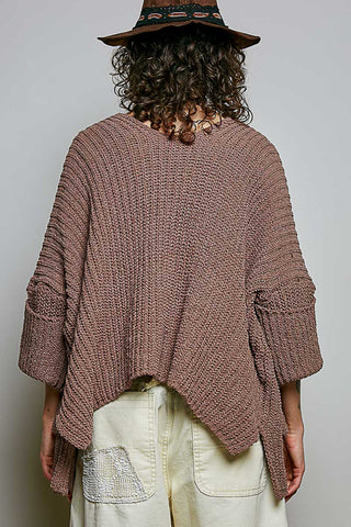 A woman wearing a chunky brown knit cardigan over a cream lace camisole and cream floral pants, photographed against a plain light gray studio background.