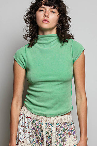 A woman wearing a bright green short-sleeve mock neck top and a floral print skirt, standing against a plain gray studio background.