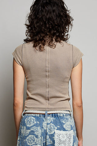 Back view of a woman wearing a beige short-sleeve knit top with a center seam and blue floral print pants against a plain gray background.