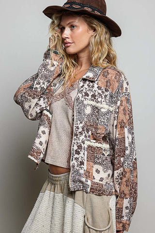 A woman wearing a bohemian-style patchwork jacket with brown, beige, and white patterns, a brown wide-brim hat, and a beige top and skirt, posed against a neutral gray background.