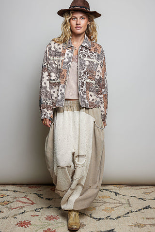 A woman modeling a bohemian-style outfit featuring a brown patterned jacket, a beige knit top, wide-leg beige pants, and a brown hat, standing against a neutral gray wall.