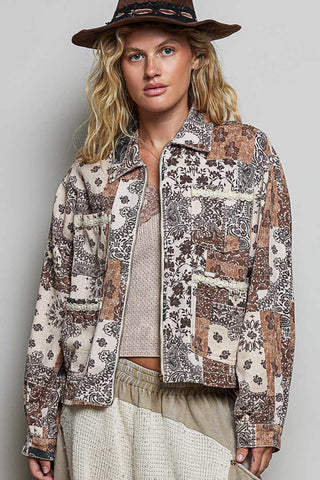 A woman wearing a cropped patchwork jacket with a beige, brown, and black floral paisley print, styled with a brown wide-brim hat, a beige knit top, and beige textured pants against a neutral gray background.