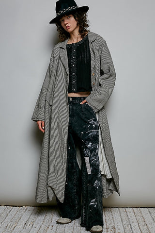 A model wearing a long gray and black striped duster coat, a black cropped knit top, dark wide-leg pants with a distressed pattern, and a black wide-brim hat, standing against a plain light gray wall.