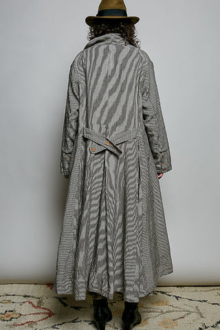 A person viewed from behind wearing a long gray and white vertically striped trench coat, a brown hat, and black boots, standing against a plain light gray wall.