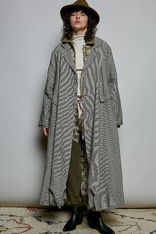 A model wearing a long gray and white wavy-striped coat over a cream turtleneck, olive green patterned pants, black boots, and a brown wide-brim hat, standing against a plain light gray wall.