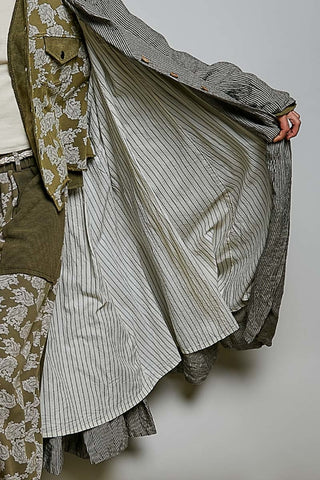 A close-up of a person wearing layered vintage-style clothing, featuring a striped shirt, a floral jacket, and olive green shorts, with a hand holding open a gray striped garment against a plain light background.