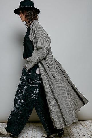 A model in profile wearing a long gray herringbone-patterned cardigan, a black hat, a black top, and dark patterned pants, walking against a light gray wall.