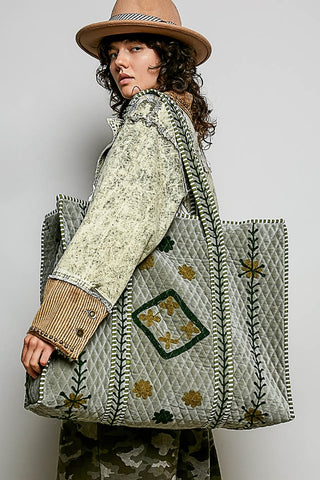 A model wearing a wide-brimmed brown hat and a patterned jacket carries a large quilted tote bag. The bag is light sage green with embroidered floral details and a diamond-shaped patch on the front pocket, set against a plain light gray studio background.