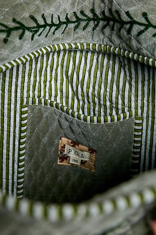 Close-up of the interior of a quilted bag or textile item, showing a gray quilted pocket with a small sewn label reading POL ONE, surrounded by green and white striped fabric and dark green embroidered stitching.